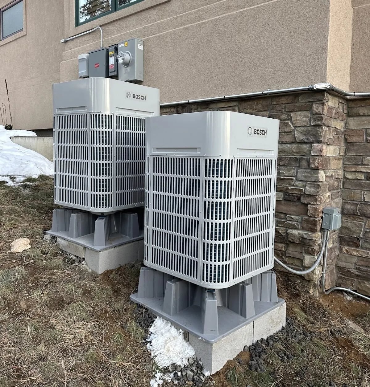 Two Bosch heat pumps on risers beside a stone wall, with snow on the ground