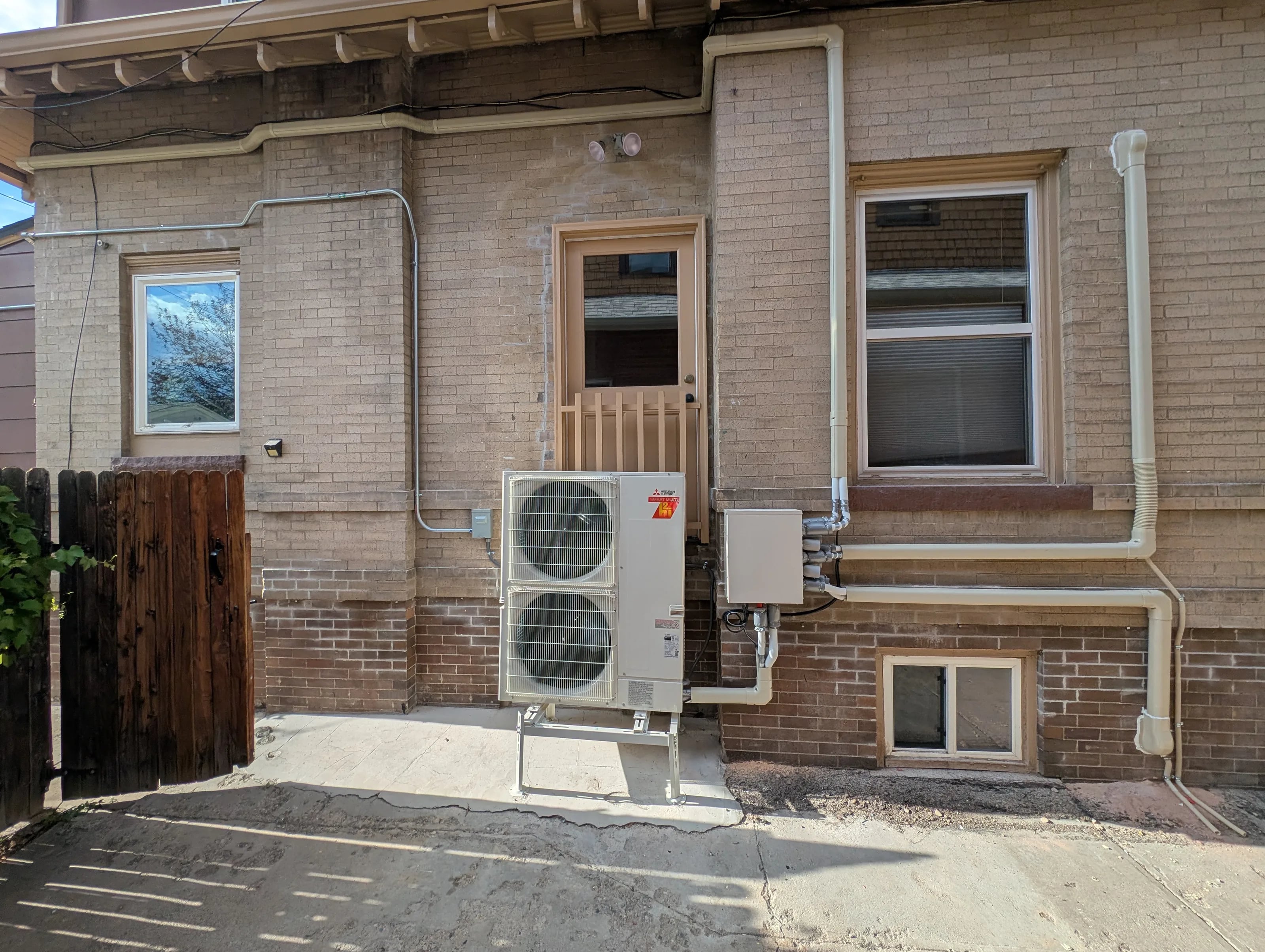 A Mitsubishi heat pump on a stand behind a tan brick house, with line set covers running to a branch box