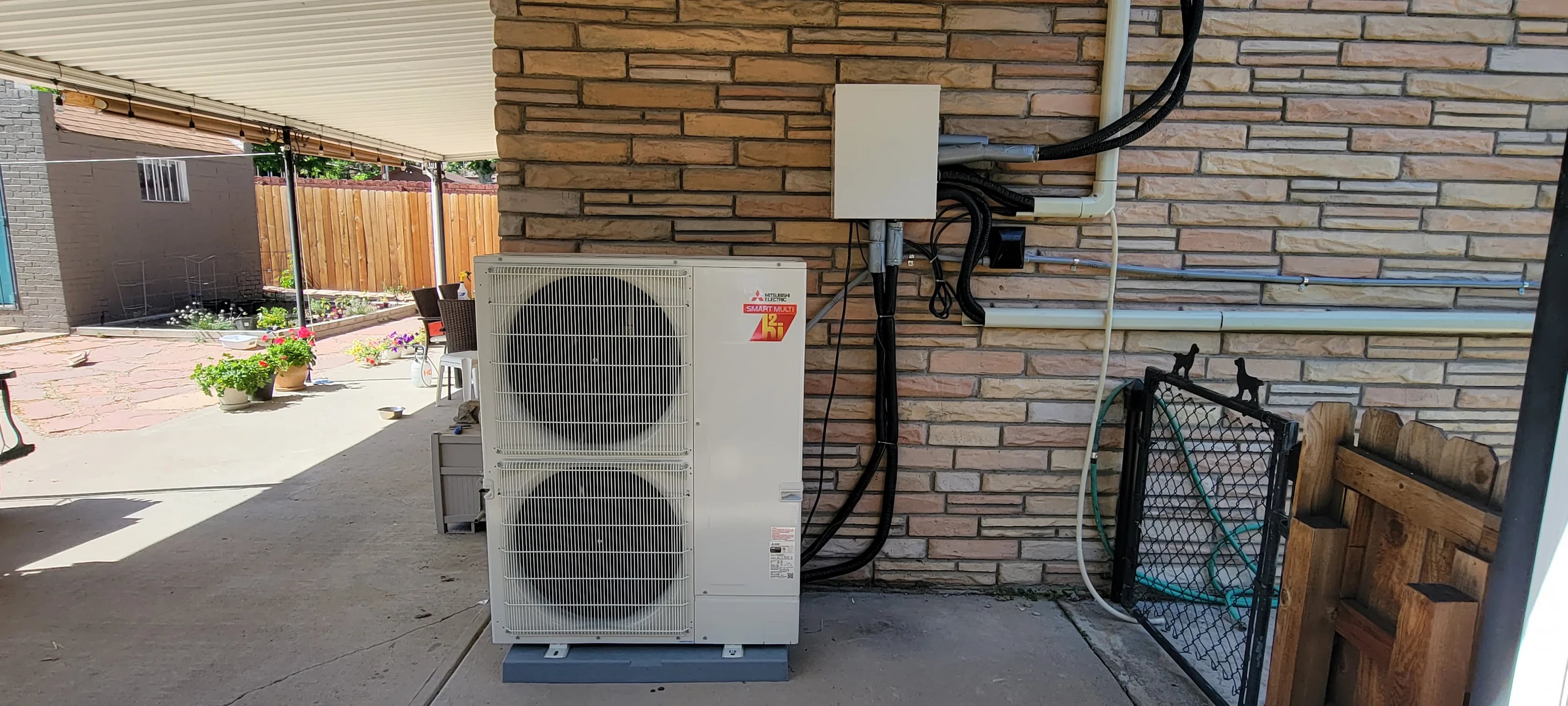 A Mitsubishi heat pump on a pad under a patio roof, with a branch box on the stone wall